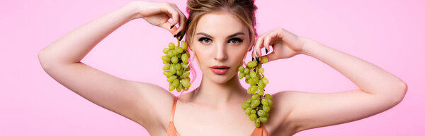 элегантная красивая блондинка держит зеленый виноград возле лица изолированы на розовый, панорамный снимок