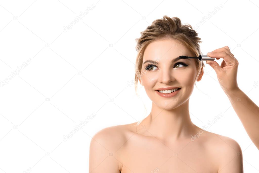 hand with brow mascara near smiling naked beautiful blonde woman isolated on white