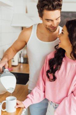 Yakışıklı adam bardağa su dolduruyor ve mutfaktaki güzel kız arkadaşına bakıyor. 