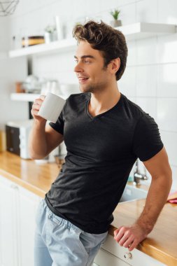 Genç adam elinde bir fincan kahve tutuyor ve mutfakta atölyenin yanında gülümsüyor. 