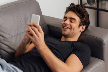 Evdeki kanepede uzanmış akıllı telefondan konuşan gülümseyen adamın seçici odağı. 