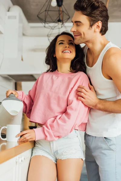 Jovem Abraçando Sorridente Mulher Derramando Água Xícara Cozinha — Fotografia de Stock