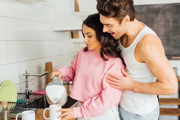 Hombre Positivo Abrazando Atractiva Mujer Vertiendo Agua Tetera Taza Encimera — Foto de Stock