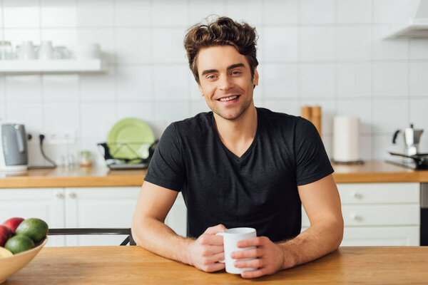 Selective focus of young man smiling at camera while holding coffee cup at table in kitchen 