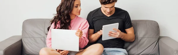 数码平板电脑和笔记本电脑在沙发上灰色背景下的年轻夫妇全景定位 — 图库照片