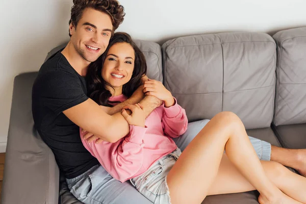 Young Couple Smiling Camera Embracing Couch Home — Stock Photo, Image