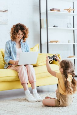 KYIV, UKRAINE - 19 Haziran 2020: Çocuk yerde otururken ve joystick tutarken dizüstü bilgisayarda çalışan genç bebek bakıcısı