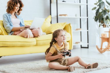 KYIV, UKRAINE - 19 Haziran 2020: Sevimli bir çocuk elinde joystick ile yerde otururken genç dadı kanepede dizüstü bilgisayar kullanıyor.