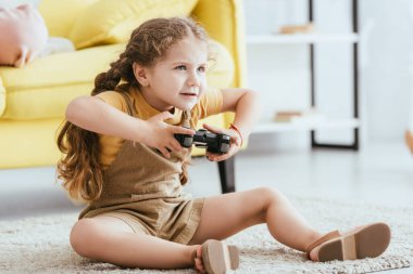 KYIV, UKRAINE - JUNE 19, 2020: adorable, focused child playing video game while sitting on floor clipart
