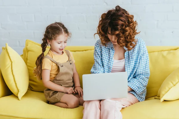 ラップトップで働いている若いベビーシッターの近くに座っているかわいい子供 — ストック写真