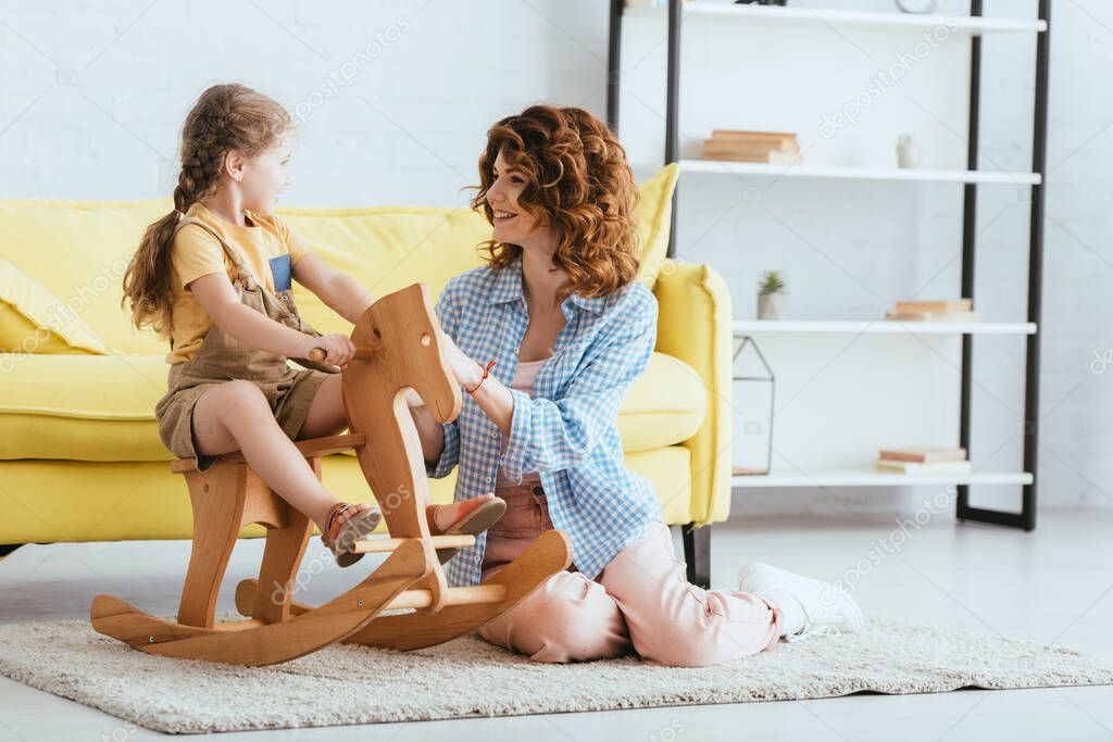smiling nanny sitting on floor and touching adorable kid riding rocking horse
