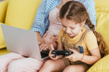 KYIV, UKRAINE - 19 Haziran 2020: sevimli çocuk bilgisayar oyunu oynarken bebek bakıcısının laptopta çalışmasını kırpmak