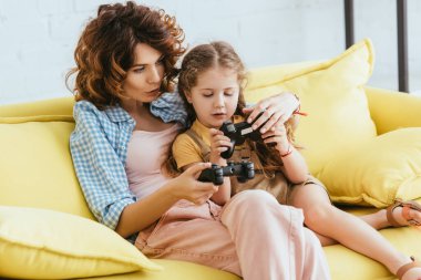 KYIV, UKRAINE - 19 Haziran 2020: genç bakıcı ve sevimli çocuk ellerinde joysticklerle sarı kanepede oturuyor.