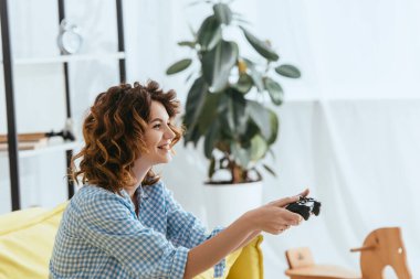 KYIV, UKRAINE - JUNE 19, 2020: side view of happy woman playing video game with gamepad clipart