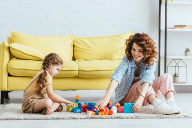 Gülümseyen bebek bakıcısı ve şirin çocuk sarı kanepenin yanında otururken yapı taşlarıyla oynuyorlar.