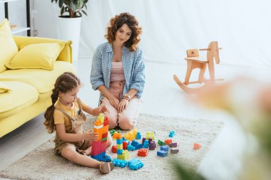 selective focus of beautiful nanny near adorable child playing with building blocks on floor  clipart
