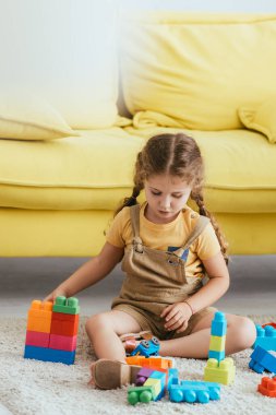 Sevimli çocuk sarı kanepenin yanında otururken yapı taşlarıyla oynuyor. 