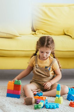 cute child playing with building blocks while sitting on floor near yellow sofa  clipart