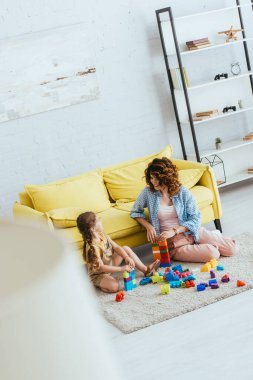 selective focus of nanny and adorable child playing with multicolored building blocks on floor in living room, high angle view clipart