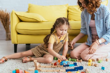 young nanny sitting near adorable kid playing with toy car near multicolored blocks on floor clipart