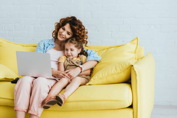 smiling nanny and child sitting on sofa and using laptop together
