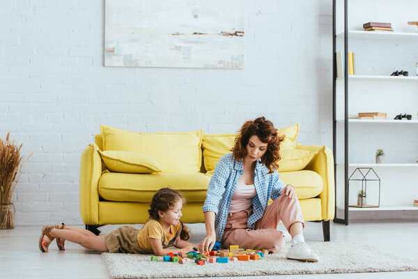 beautiful nanny and cute child playing with multicolored blocks on floor near yellow sofa