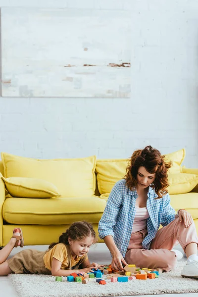 年轻的保姆和可爱的孩子在黄色沙发附近的地板上玩五颜六色的方块 — 图库照片
