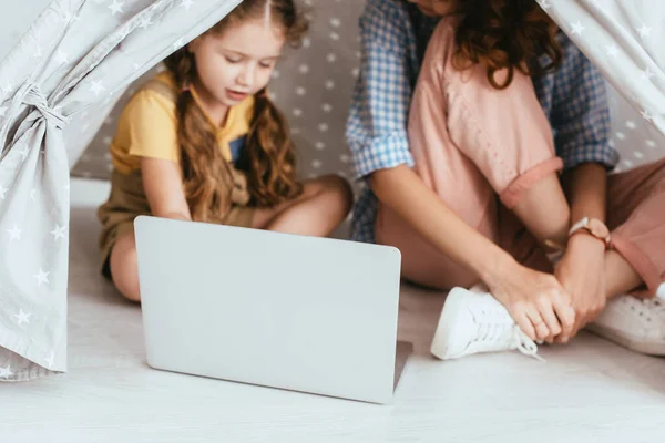 おもちゃのウィグワムでノートパソコンを使ってかわいい子供の近くに座っているベビーシッターのクロップビュー 選択的フォーカス — ストック写真