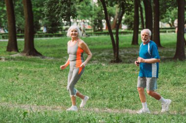 Positive senior coupe looking at camera while jogging in park  clipart