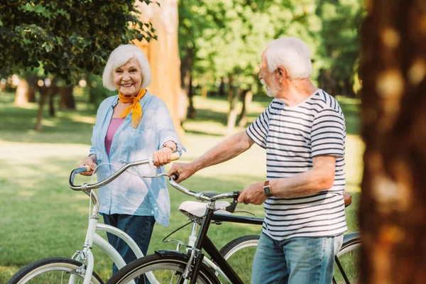 Selective Focus Smiling Senior Woman Bicycle Looking Camera Husband Park — Stock Photo, Image