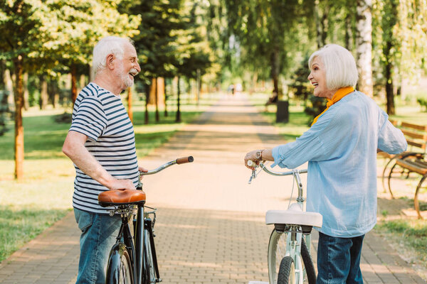 Senior woman looking at smiling husband while standing near bicycle in park 