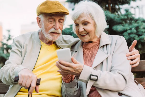 Parktaki Bankta Kocasının Yanında Akıllı Telefon Tutan Gülümseyen Yaşlı Kadının — Stok fotoğraf