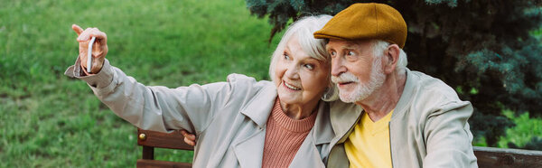 Panoramic shot of smiling senior woman taking selfie with smartphone near husband on bench in park 