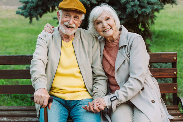 Grey haired couple holding hands and smiling at camera on bench in park 