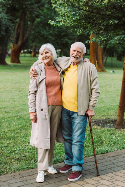 Senior man smiling at camera and hugging cheerful wife on walkway in park 