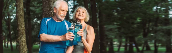 Panoramic shot of smiling senior woman holding sports bottle near husband in park