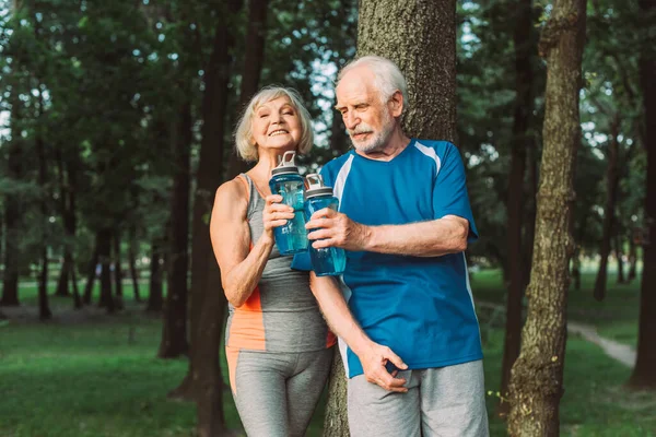 在公园靠近丈夫和树的地方拿着运动瓶微笑的老年妇女 — 图库照片