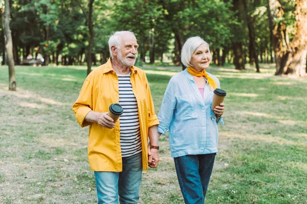 公園を歩いている間に行くためにコーヒーを持っている高齢者のカップルを笑顔 — ストック写真