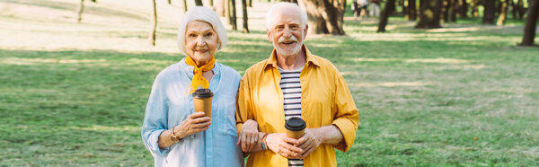 Horizontal crop of smiling senior couple with coffee to go looking at camera in park 