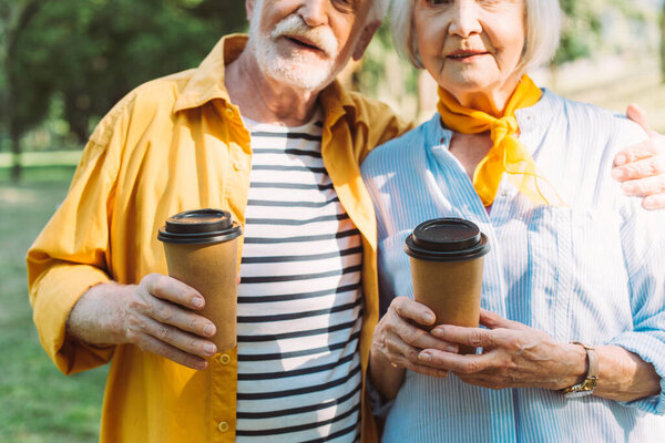 Cropped view of senior man holding coffee to go and embracing wife in park 
