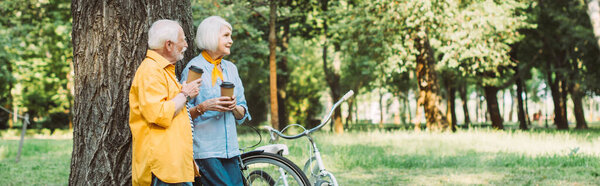 Панорамный снимок улыбающейся пожилой женщины с одноразовой чашкой рядом с мужем и велосипедами в парке 