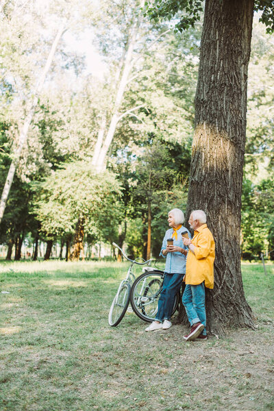 Senior man with paper cup looking at smiling wife near tree and bikes in park 