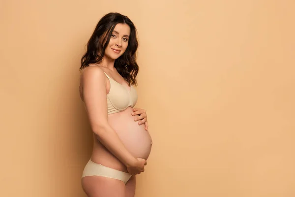 ブルネット妊娠中の女性で下着タッチ腹と見ますカメラオンベージュ — ストック写真