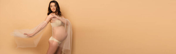 Pregnant woman in underwear and chiffon sleeves posing on beige, horizontal image
