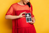Teilaufnahme einer schwangeren Frau in rotem Outfit mit Ultraschalluntersuchung in Bauchnähe auf gelb