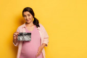 Schwangere zeigt Ultraschallbild, während sie Bauch auf gelb berührt  