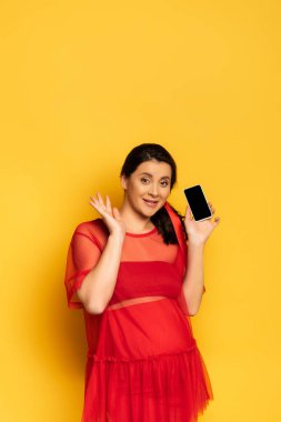 Esmer, hamile bir kadın, sarı ekranda boş ekranlı akıllı telefonu gösterirken el sallıyor.