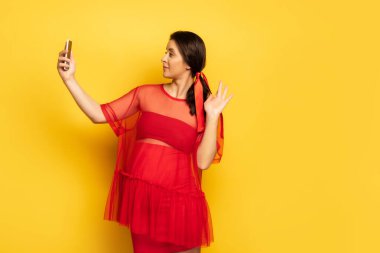 Genç hamile bir kadın, kırmızı tunik giyip sarı telefonda görüntülü konuşma yaparken el sallıyor.