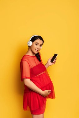Kablosuz kulaklıklı hamile bir kadın elinde boş ekranlı akıllı bir telefon ve sarı karınına dokunuyor.