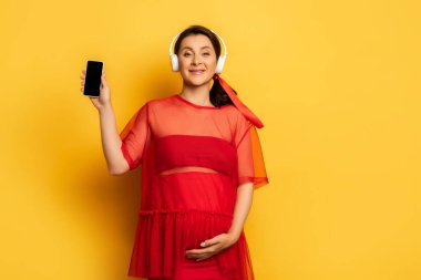 Kablosuz kulaklık takan hamile bir kadın sarı karnına dokunurken akıllı telefon gösteriyor.